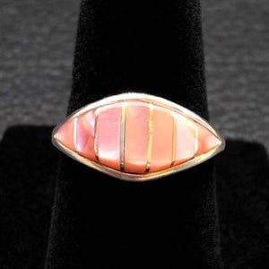 May include: A silver ring with an oval-shaped, inlaid design. The inlay features alternating vertical stripes of pink and peach-colored material, separated by thin gold-colored metal strips. The ring is displayed on a black surface.