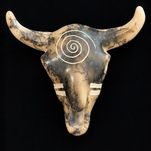 May include: A ceramic bull skull wall hanging featuring a smooth cream-colored surface with dark gray smoke-fired marbling patterns. The skull is decorated with a prominent white etched spiral on the forehead and two horizontal white stripes on each cheek.