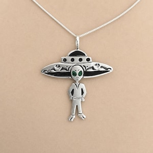 May include: A silver pendant necklace featuring an alien figure standing beneath a large, detailed UFO saucer. The alien has two small green gemstone eyes, and the saucer includes black enamel accents and circular patterns on its metallic surface.