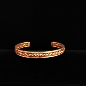 May include: A copper cuff bracelet featuring a central twisted rope design flanked by two smooth, thin bands. The metal has a polished, warm metallic finish with an open back, showcasing a continuous circular structure with rounded ends.