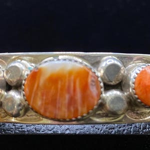 May include: A silver cuff bracelet featuring three oval-shaped orange stones set in metal bezels. The central stone is larger than the two flanking stones. The silver band is decorated with small metal spheres and leaf-like engravings.
