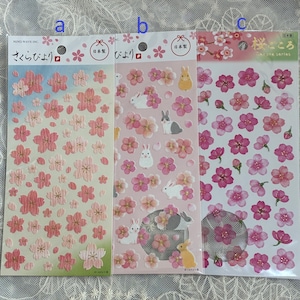 以下が含まれることがあります： 桜の花のデザインが施された3枚のステッカーシート。1枚はグラデーションの背景にピンクの花、もう1枚はピンクの花と白いウサギ、3枚目はピンクの花。日本語のテキストが表示されています。
