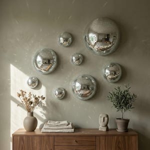 Op de afbeelding: Een verzameling zilveren discobol wanddecoraties in verschillende maten, gemonteerd op een neutrale muur. De reflecterende oppervlakken creëren een sprankelend effect. Een houten kast eronder bevat decoratieve items.