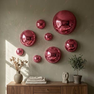 May include: A display of pink disco balls of different sizes fixed to a wall. The mirrored surfaces reflect light, creating a sparkling effect. A wooden cabinet below holds decorative items, including a vase with dried flowers and a small potted plant.