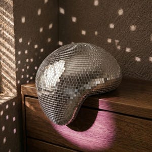 Op de afbeelding: Een afgerond, asymmetrisch decoratief object bedekt met kleine, reflecterende zilveren spiegeltjes. Het oppervlak bestaat uit een raster van vierkante glazen stukjes die het licht opvangen en verspreiden, wat een glinsterend effect geeft.