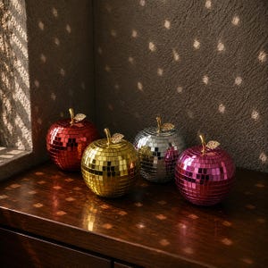 Könnte beinhalten: Vier apfelförmige Ornamente, die mit reflektierenden Spiegelkacheln in Rot, Gold, Silber und Pink bedeckt sind. Jeder Apfel hat einen kleinen, metallisch goldenen Stiel mit einem einzelnen Blatt an der Oberseite, was für ein funkelndes Aussehen sorgt.