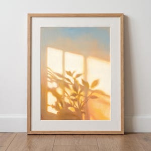 Peut inclure: Une impression d'art encadrée représentant une peinture de lumière dorée traversant une fenêtre, projetant des ombres de feuillage sur un mur. L'œuvre est insérée dans un cadre en bois clair avec une bordure passe-partout blanche.