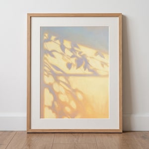 May include: A framed wall art print featuring a painting of soft, golden sunlight casting elongated, purple-toned shadows of leaves and branches across a surface, enclosed in a light-colored wooden frame with a white mat border.