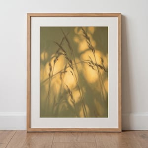 Könnte beinhalten: Ein gerahmter Kunstdruck mit einem weich gezeichneten, goldfarbenen Bild von hohem Gras. Das Kunstwerk befindet sich in einem hellen Holzrahmen mit weißem Passepartout und zeigt eine warme, sonnendurchflutete Ästhetik mit verschwommenen Silhouetten.
