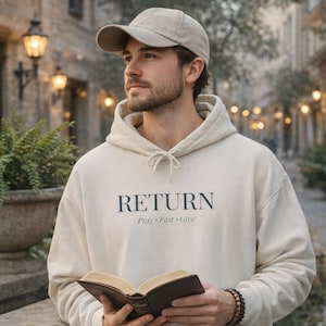 Może przedstawiać: Beżowa bluza z kapturem z napisem "RETURN" w kolorze granatowym, z napisem "Pray • Fast • Give" poniżej. Osoba na zdjęciu ma na sobie beżową czapkę z daszkiem i trzyma książkę.
