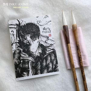 Puede incluir: Ilustración en blanco y negro de un personaje con el texto "Guts" en una tarjeta blanca. Dos pinceles de caligrafía con mangos marrones y beige y puntas blancas descansan sobre una tela rosa. El texto "INKU ANIME" y "INKU ART STUDIO" está en la parte superior.