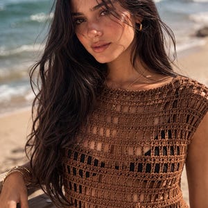 May include: A brown crochet top with a square pattern. The top is sleeveless and has a scoop neckline. The background shows a beach with the ocean in the distance. The top is worn over a black top.
