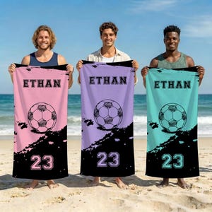 May include: Three rectangular beach towels featuring a soccer ball graphic, the name ETHAN, and the number 23. The towels are pink, light purple, and teal, each with a black bottom section featuring a distressed paint splatter design.
