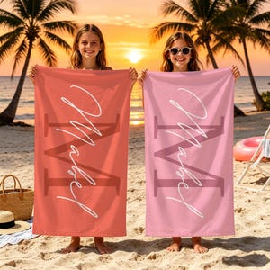 Può includere: Due teli mare rettangolari, uno color corallo e uno rosa chiaro, ciascuno caratterizzato da una grande lettera M scura sullo sfondo e dal nome Mabel scritto in un carattere corsivo bianco al centro della superficie in tessuto.