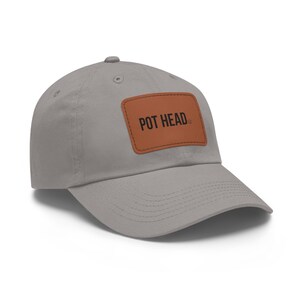 May include: A grey baseball cap with a curved peak. The cap has a brown leather patch with the words "POT HEAD" printed in black. The cap is made of a soft fabric and has a button on top.