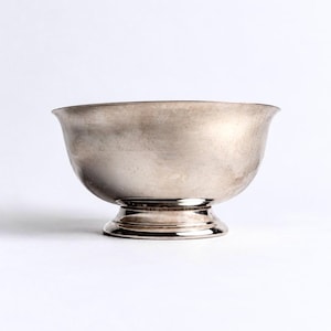 May include: A polished silver-toned metal bowl with a smooth, curved exterior and a wide, flared rim. The bowl features a circular pedestal base that elevates the main body, showcasing a reflective, metallic finish with subtle surface variations.