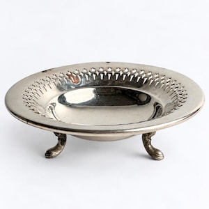 May include: A round silver-toned metal dish featuring a wide, flat rim with a decorative pierced lattice pattern. The bowl has a smooth, reflective interior basin and is supported by three small, curved feet with detailed claw-like ends.