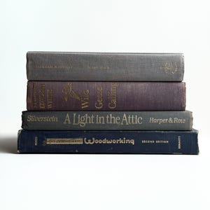 May include: A stack of four hardcover books. The top book is grey with Moby Dick by Herman Melville. The second is purple with Wild Geese Calling by Edward White. The third is grey with A Light in the Attic by Silverstein. The bottom is blue Industrial Arts Woodworking.