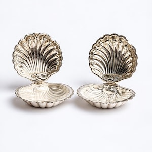 May include: Two open silver-toned clam shell dishes with a polished, reflective finish. Each dish features a hinged design with scalloped edges and deep, radiating grooves that mimic the natural texture of a seashell, creating a metallic look.