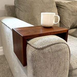 May include: A dark brown wooden sofa arm tray with a white mug on top. The tray is rectangular and fits over the arm of a light gray sofa. The mug is white and has a handle.