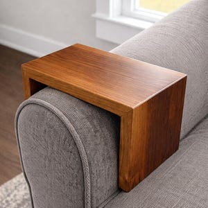 May include: A rectangular wooden sofa arm table in a warm brown finish. The table is designed to rest on the arm of a gray upholstered sofa. The wood grain is visible, and the table has a clean, modern design.