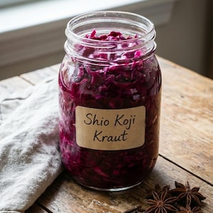 Pode incluir: Um frasco de vidro cheio de repolho roxo desfiado vibrante, rotulado como "Shio Koji Kraut". O frasco está sobre uma superfície de madeira, acompanhado de anis estrelado e cravos. Um pano cinza é parcialmente visível.