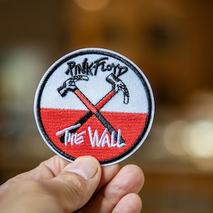 以下が含まれることがあります： ピンク・フロイドのロゴが入った刺繍パッチ。交差したハンマーと「THE WALL」の文字が特徴です。パッチは円形で、白い縁取り、赤い下部、黒と白の刺繍が施されています。