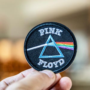 May include: A round, black embroidered patch featuring the words "PINK FLOYD" in white, arched text. A blue triangle is in the center, with a white line extending from the left and a rainbow on the right. The patch is held in a hand.
