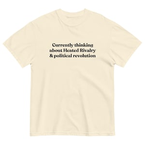 May include: Cream-colored t-shirt with the text "Currently thinking about Heated Rivalry & political revolution" in black. The shirt has a crew neck and short sleeves. The fabric appears to be a soft cotton blend.