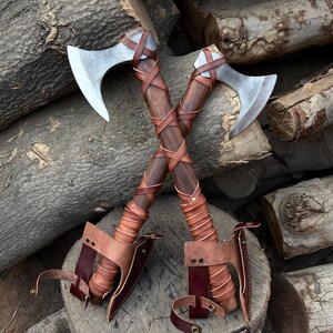 May include: Two crossed axes with polished steel blades and dark wooden handles wrapped in brown leather. The axes rest on a wooden surface against a backdrop of stacked logs. Each axe has a leather sheath.