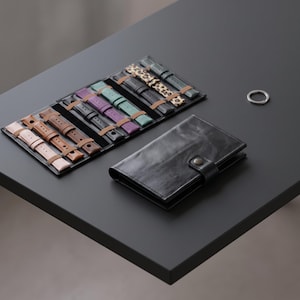 May include: A black leather watch strap organiser case with a snap closure, containing multiple watch bands in various colours including pink, brown, black, purple, teal, and leopard print, alongside a small silver metal ring.