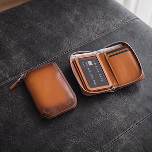 May include: Two tan leather wallets with silver-tone metal zips. One wallet is closed, while the other is open, revealing interior card slots, a clear identification window, and a compartment containing a paper note and a black premium card.