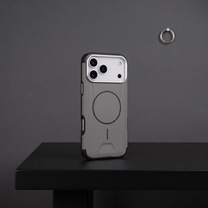 Puede incluir: Una funda de smartphone gris con un marco de módulo de cámara plateado y un anillo de carga magnético circular en la parte posterior. La funda presenta un acabado mate, un detalle de línea vertical cerca de la base y bordes reforzados.
