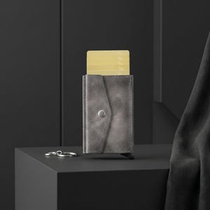 May include: A grey leather card holder wallet with a triangular flap closure secured by a silver metal snap button. The wallet features a vertical design with a single light yellow card partially extended from the top and a silver metal key ring.