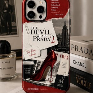 May include: A red iPhone case featuring a collage of fashion-related imagery. The case includes the text "The Devil Wears Prada 2", a red high heel, and quotes about fashion. The case also has a black camera bump.