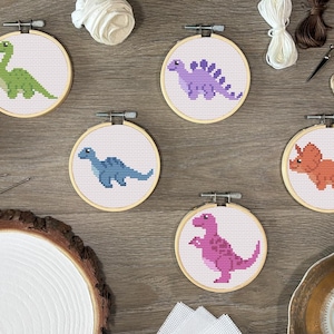 May include: Five circular wooden embroidery hoops containing cross-stitch patterns of dinosaurs. The designs feature a green long-necked dinosaur, a purple stegosaurus, a blue dinosaur, an orange triceratops, and a pink tyrannosaurus rex.