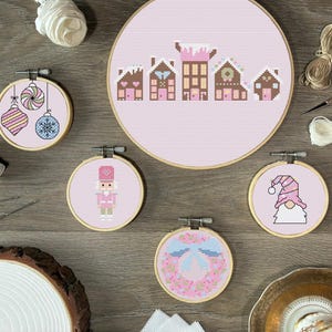以下が含まれることがあります： 薄いピンク色の布にクロスステッチが施された5つの木製刺繍枠。ジンジャーブレッドハウスの列、3つのクリスマスオーナメント、くるみ割り人形、縞模様の帽子のノーム、青いリボンの付いた花のリースがデザインされています。