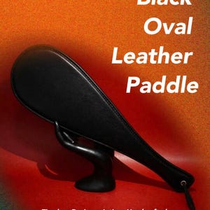 May include: A black oval leather paddle with visible stitching along the edges and a small metal eyelet at the end of the handle. The text "Black Oval Leather Paddle" and "Timeless Design • Artisan Handcrafted" is printed on the image.