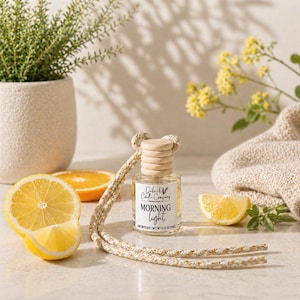 May include: A car air freshener with a wooden cap and a white and gold cord. The label reads "Morning Light." The image includes a sliced lemon, an orange slice, and a small vase with greenery.