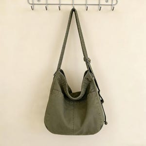Puede incluir: Bolso de hombro de lona verde oliva con correa ajustable. El bolso tiene un compartimento principal grande y dos bolsillos exteriores. Cuelga de un perchero de metal en una pared color crema.