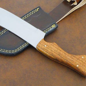 May include: A fixed-blade knife with a silver-toned metal blade and a contoured wooden handle secured by two small metal rivets. The knife is accompanied by a dark brown leather sheath featuring visible yellow stitching along the edges.