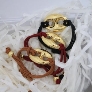 Puede incluir: Pulseras de metal dorado con diseño en forma de frijol, combinadas con cuerdas negras, marrones y rojas. Las pulseras se exhiben sobre papel blanco. La joyería tiene una estética moderna y minimalista.