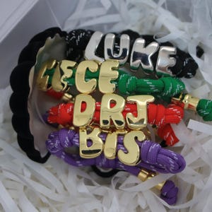 Könnte beinhalten: Eine Sammlung personalisierter Armbänder mit Namen und Initialen. Die Armbänder verfügen über farbenfrohe geflochtene Kordeln in Schwarz, Grün, Rot und Lila, die mit goldenen und silbernen Buchstabenanhängern mit Namen und Initialen verziert sind. Die Armbänder werden in einer weißen Schachtel mit geschreddertem Papier präsentiert.