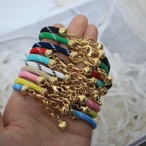 Könnte beinhalten: Eine Sammlung farbenfroher Seilarmbänder mit goldfarbenen Verschlüssen und herzförmigen Anhängern. Die Armbänder weisen eine Vielzahl von Farben auf, darunter Schwarz, Grün, Marineblau, Rot, Türkis, Weiß, Rosa, Gelb und Blau. Jedes Armband ist mit goldfarbenen Metallakzenten verziert.