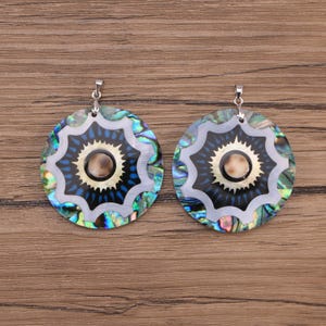 May include: A pair of circular pendants featuring an iridescent abalone shell outer ring, a white scalloped border, and a central black starburst pattern with a small, round, tan-colored inlay. Each pendant includes a silver-toned metal bail.