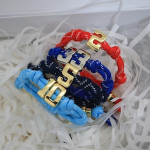 Può includere: Quattro braccialetti in corda intrecciata rossa, blu e azzurra, ciascuno con una chiusura in metallo color oro che mostra un numero. I braccialetti sono immersi in carta bianca. I numeri sulle chiusure sono 10, 33 e 2.