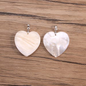 May include: Two heart-shaped pendants made of iridescent shell material. Each pendant features a smooth, polished surface with natural swirling patterns in shades of cream and off-white, and includes a silver-toned metal bail at the top.
