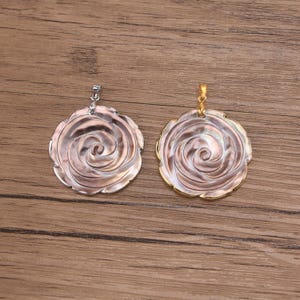 May include: Two circular pendants featuring a carved iridescent shell rose design. One pendant has a silver-toned metal bail, while the other features a gold-toned metal bail. Both pieces display a swirling floral pattern with a pearlescent finish.
