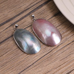 Puede incluir: Dos colgantes de forma ovalada con superficies lisas e iridiscentes en monturas de metal plateado. Un colgante muestra un tono gris carbón oscuro con sutiles reflejos brillantes, mientras que el otro presenta un tono rosa pálido con lustre nacarado.