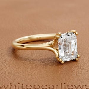 Pode incluir: Um anel de noivado de ouro com um diamante de corte radiante. O anel apresenta um diamante claro em forma retangular preso por quatro garras. O texto "whitepearljewels" é visível.